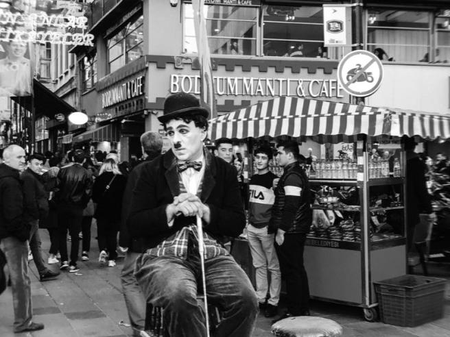 black and white photo of a Charlie Chaplin actor sitting on a high chair observing a busy street
